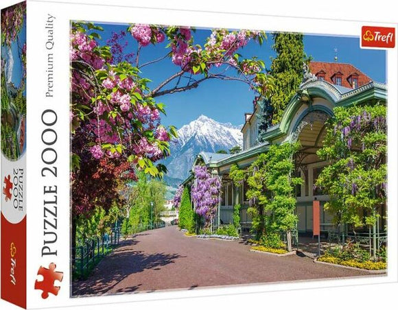 Trefl jigsaw puzzle featuring a picturesque scene from Merano, Italy. The image showcases vibrant floral trees, a charming pathway, and snow-capped mountains in the background, all within a beautiful spring setting. The puzzle contains 2000 pieces, designed for adults and puzzle enthusiasts seeking a scenic challenge.