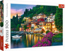 Trefl jigsaw puzzle featuring a scenic view of Lake Como, Italy. The image depicts colorful houses along the shoreline amidst lush greenery and vibrant flowers, with a calm lake and a small boat reflecting the evening sky.