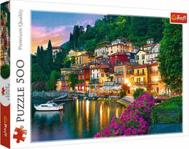 Trefl jigsaw puzzle featuring a scenic view of Lake Como, Italy. The image depicts colorful houses along the shoreline amidst lush greenery and vibrant flowers, with a calm lake and a small boat reflecting the evening sky.
