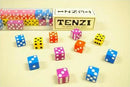 A colorful collection of dice in various colors including blue, pink, yellow, and green scattered on a light background. The game box labeled 'TENZI' is visible in the top center, showcasing the game as 'the world's fastest game'. Each die features unique dot patterns, enhancing the vibrant display and inviting gameplay.