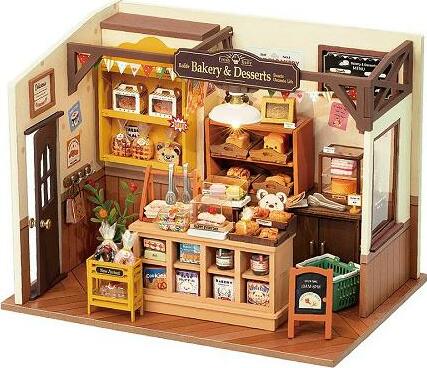 Becka's Baking House DIY kit from Robotime, showcasing a miniature bakery scene complete with shelves of baked goods, decorative items, and a warm, inviting atmosphere. The model features a sign reading 'Bakery & Desserts', a front counter filled with pastries, and charming details like teddy bears and plant decorations.