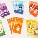 A colorful assortment of game cards from Kinoko by Helvetiq. The cards feature whimsical illustrated characters like a snowman in a top hat, vibrant mushrooms, and a singing snow figure, showcasing numbers prominently at the top. The design is playful and perfect for family-friendly games.