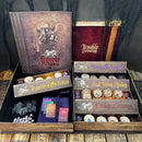 A detailed view of the game 'Blood on the Clocktower' by Pandemonium Institute. The image shows the game box open, displaying compartments filled with colorful tokens, cards, and character boards. The design features dark, immersive artwork with the title prominently displayed, alongside sections labeled 'Travellers & Fabled', 'Sects & Violets', and 'Had Aborn Rising'. The overall aesthetic is mysterious and captivating, perfect for game enthusiasts.