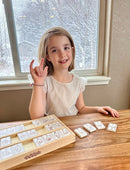 A young girl with short brown hair sits at a wooden table, smiling and holding her hand in a sign language gesture. In front of her are wooden tiles featuring illustrations of letters from the sign language alphabet arranged on a tray. The background shows a snowy landscape through a window, creating a cozy atmosphere.