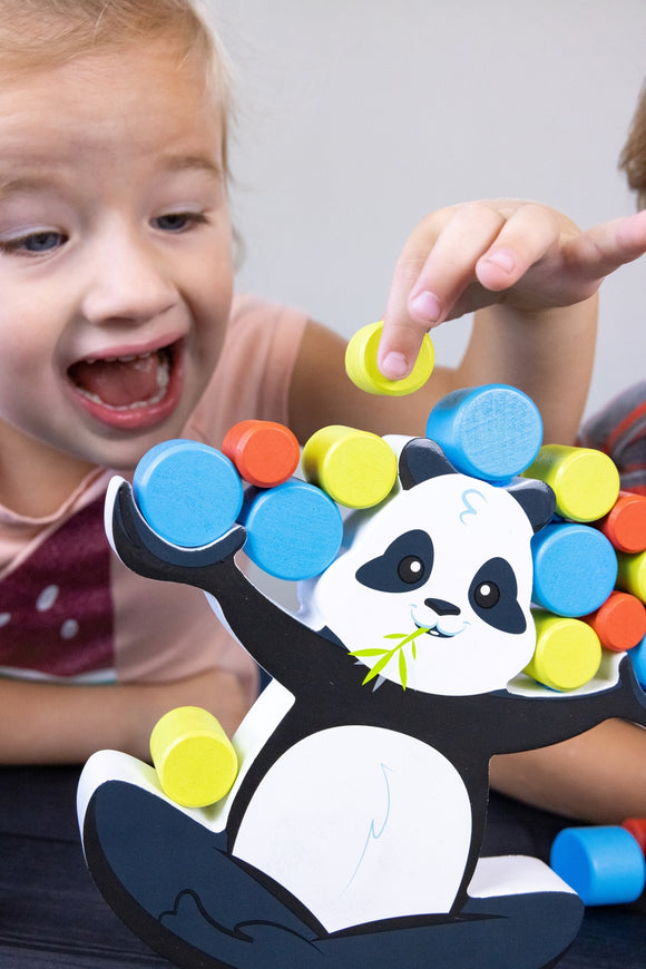 A child joyfully plays with BoomBoom the Balancing Panda game. The colorful wooden pieces are being stacked on the smiling panda figure, engaging the child's focus and excitement as they balance the shapes.