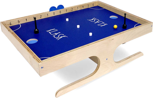 Klask game from Buffalo Games featuring a wooden board with a vibrant blue playing surface. The board has two black player pieces, a yellow ball, and several white pieces scattered on it, along with scorekeeping markings along one edge.