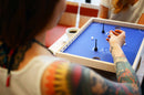 A close-up view of two people playing Klask, a dynamic tabletop game by Buffalo Games. One player is poised to make a move, with a focus on the blue playing surface featuring small pieces and black poles. A cup of coffee is visible in the background.