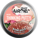A round tin of Crazy Aaron's Mini Thinking Putty in Rose Gold color. The lid features the brand's logo and product name, with a shimmering putty displayed underneath. The background includes hints of pink roses and pastel colors, emphasizing the playful and creative nature of the product. Suitable for ages 3 and up.