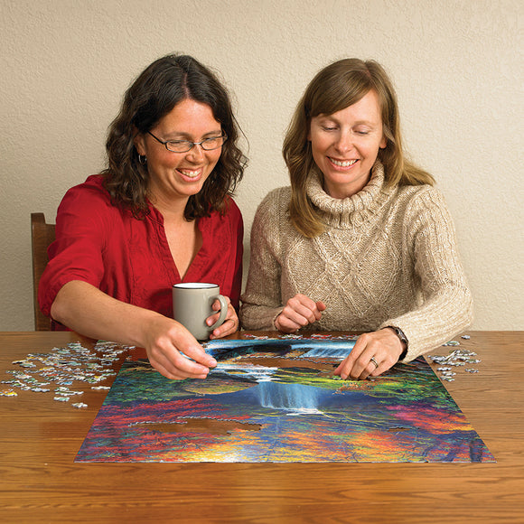 Two women enjoying a jigsaw puzzle titled 'Mystic Falls in Autumn' by Cobble Hill. They are seated at a table surrounded by puzzle pieces, laughing and working together on the vibrant autumn scene featuring a waterfall and colorful foliage, with one woman holding a coffee mug.