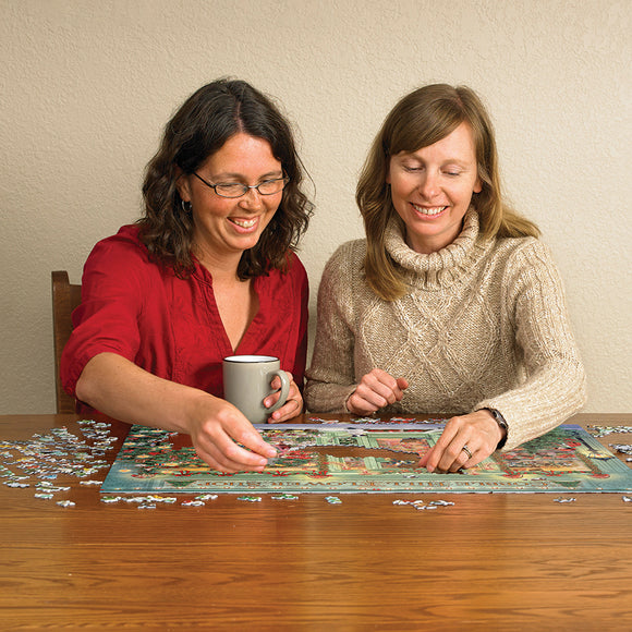 Two women enjoying a jigsaw puzzle titled 'Christmas Flower Shop' by Cobble Hill. They are seated at a wooden table, one holding a mug while both focus on assembling the colorful pieces of the puzzle featuring vibrant flowers and a festive scene.