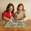 Two women smiling and collaborating on The Purrfect Bookshelf jigsaw puzzle. One woman holds a coffee mug while the other places a puzzle piece, surrounded by partially completed sections and scattered puzzle pieces on a wooden table.