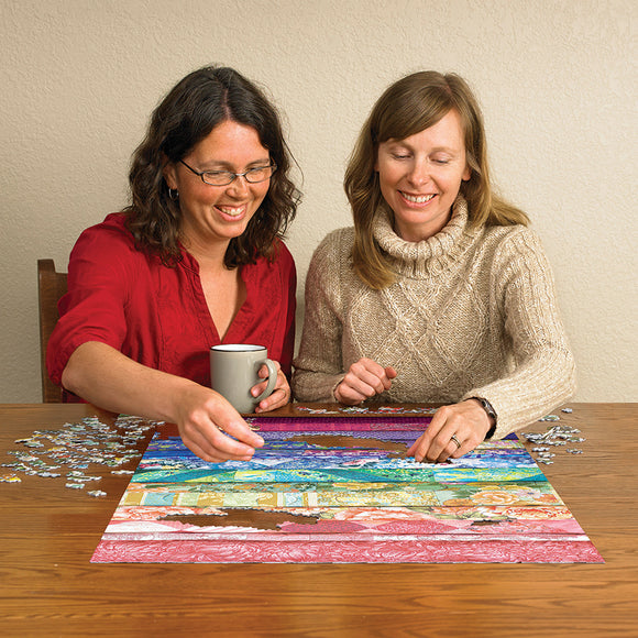 Two women enjoying a jigsaw puzzle titled 'Comfortable Rainbow' by Cobble Hill. They are piecing together a colorful puzzle on a wooden table. One woman holds a coffee mug while the other works on connecting pieces of vibrant sections.