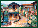 A vibrant jigsaw puzzle titled "Fountain on the Square" by Cobble Hill. The image captures a picturesque cobblestone street scene with a fountain at the center, charming buildings adorned with colorful flowers, a quaint café sign, and a scenic landscape in the background. Ideal for puzzlers seeking a delightful challenge and beautiful artwork.