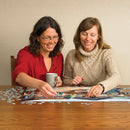 Two women joyfully assembling the 'Fountain on the Square' jigsaw puzzle from Cobble Hill. One woman is holding a mug while placing a puzzle piece, and the other is smiling as she helps. The puzzle features vibrant artwork depicting a fountain and surrounding scenery, with pieces scattered around them on a wooden table.