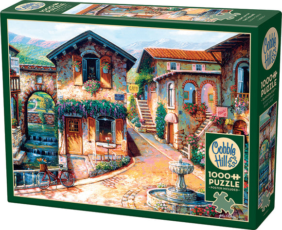 Cobble Hill jigsaw puzzle titled 'Fountain on the Square', featuring a charming village scene with cobblestone pathways, a central fountain, lush greenery, and a café. The puzzle contains 1000 pieces and includes a poster, showcasing vibrant details of the picturesque setting.