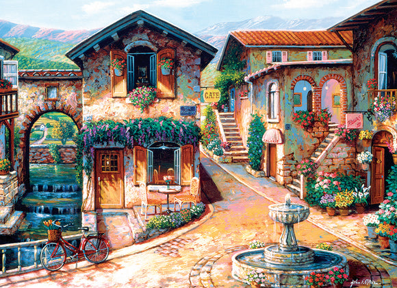 A picturesque jigsaw puzzle titled 'Fountain on the Square' by Cobble Hill, depicting a charming European square with colorful buildings, vibrant flowers, and a serene fountain. The scene captures a quaint atmosphere with outdoor seating, bicycles, and signs for a café and gallery.