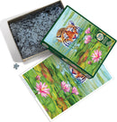 Land of the Lotus jigsaw puzzle by Cobble Hill, showcasing a beautiful scene with a tiger peeking out from a serene pond surrounded by lily pads and flowers. The box is partially open, revealing the puzzle pieces inside, which are gray and intricately shaped. The artistic image includes vibrant greens and pinks, offering a picturesque view of nature.