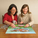 Two women sitting at a wooden table, enjoying a jigsaw puzzle titled 'Land of the Lotus' by Cobble Hill. One woman, wearing glasses and a red shirt, is placing a piece of the puzzle featuring a tiger and pink lotus flowers into the scene, while the other woman in a cozy sweater smiles and holds a mug. Surrounding them are scattered puzzle pieces, creating a warm and engaging atmosphere for puzzle enthusiasts.