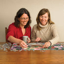 Two women joyfully assembling 'The Garden Wall' jigsaw puzzle by Cobble Hill on a wooden table, surrounded by scattered puzzle pieces. One woman holds a mug, while they focus on completing the colorful floral image.