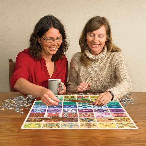 Two women are enjoying a jigsaw puzzle featuring various colorful quilt blocks. One woman is holding a mug while the other concentrates on placing a puzzle piece, with several pieces scattered around on the wooden table.