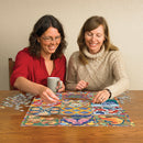 Two women enjoying a festive holiday jigsaw puzzle titled "Twelve Days of Christmas Quilt" by Cobble Hill. One woman in a red shirt is placing a puzzle piece, while the other in a cozy sweater smiles, holding a coffee mug. The colorful puzzle features intricate holiday designs spread across a wooden table, with some puzzle pieces scattered around.