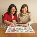 Two women enjoy completing the Cobble Hill Bicycles jigsaw puzzle together on a wooden table. One woman holds a coffee mug, while both focus on the colorful puzzle pieces depicting various bicycles laid out on the board. Surrounding the puzzle are scattered pieces, creating a warm and engaging atmosphere for a fun puzzle-solving experience.