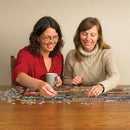 Two women joyfully assembling a Cobble Hill jigsaw puzzle on a wooden table. One woman holds a coffee mug while placing a puzzle piece, and the other smiles as she contributes to the puzzle, surrounded by scattered pieces.