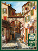 A 1000-piece jigsaw puzzle by Cobble Hill featuring a scenic street view titled 'Ceramica.' The artwork depicts charming buildings with colorful shutters and an archway. Restaurants and shops line the cobbled street, creating a warm, inviting atmosphere. A poster is included with the puzzle.