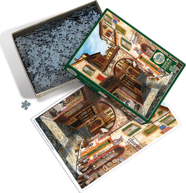 The Cobble Hill Ceramica jigsaw puzzle featuring a picturesque street scene with colorful buildings and a cobblestone path. The puzzle pieces are spread out in a box, and the box cover displays the completed image, showcasing detailed artwork and vibrant colors.