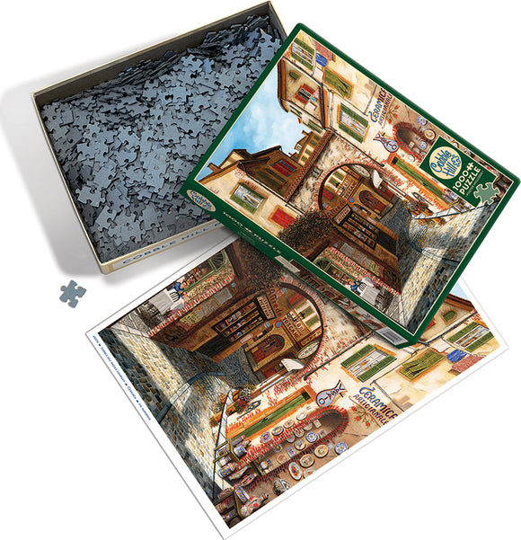 The Cobble Hill Ceramica jigsaw puzzle featuring a picturesque street scene with colorful buildings and a cobblestone path. The puzzle pieces are spread out in a box, and the box cover displays the completed image, showcasing detailed artwork and vibrant colors.