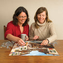 Two women engaged in assembling the Ceramica jigsaw puzzle by Cobble Hill, featuring a colorful town scene. One woman is holding a mug and placing a puzzle piece, while the other smiles and holds a piece ready to fit. The wooden table is scattered with additional puzzle pieces, creating a cozy and interactive atmosphere.