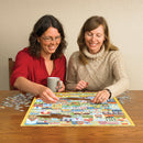 Two women enjoy assembling the 'Home Sweet Home' jigsaw puzzle by Cobble Hill, seated at a wooden table with puzzle pieces scattered around. One woman holds a coffee mug while carefully placing a puzzle piece into the vibrant, illustrated scene of a friendly neighborhood filled with colorful houses and cheerful details.