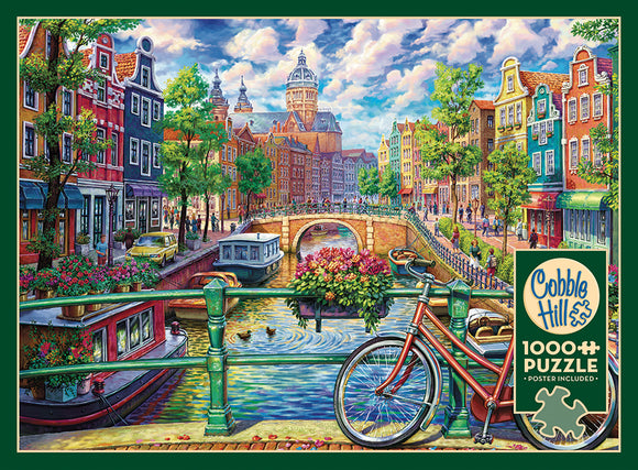 A vibrant jigsaw puzzle depicting a picturesque Amsterdam canal scene, featuring colorful buildings, a bridge, and a bicycle in the foreground. The image showcases lush greenery, boats on the water, and people enjoying the scenery, capturing the charm of the city. The puzzle includes 1000 pieces and a poster for reference, made by Cobble Hill.