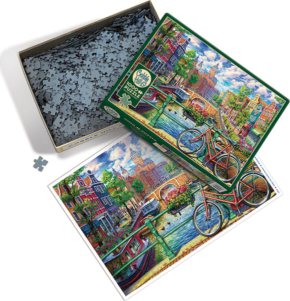 Cobble Hill 1000-piece jigsaw puzzle featuring a vibrant Amsterdam canal scene. The box is open, showcasing numerous puzzle pieces and a colorful image of bicycles by the water, charming houses, and lush greenery.