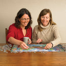 Two women enjoy assembling the Amsterdam Canal jigsaw puzzle on a wooden table, with pieces scattered around them. One woman holds a gray mug while they discuss which pieces to fit next. The colorful puzzle features a scenic depiction of Amsterdam's canals and buildings.