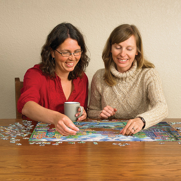Two women enjoy assembling the Amsterdam Canal jigsaw puzzle on a wooden table, with pieces scattered around them. One woman holds a gray mug while they discuss which pieces to fit next. The colorful puzzle features a scenic depiction of Amsterdam's canals and buildings.