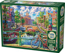 Cobble Hill jigsaw puzzle titled 'Amsterdam Canal', featuring a vibrant, colorful depiction of a canal in Amsterdam with lush greenery, quaint buildings, and a bicycle in the foreground. The box includes a 1000-piece puzzle and a decorative poster of the artwork.