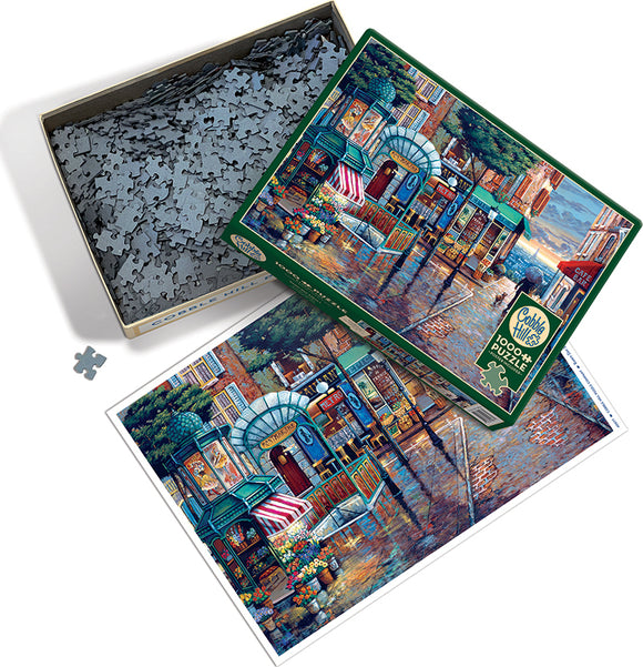 The Cobble Hill jigsaw puzzle titled "Rainy Day Stroll" features a colorful scene of a charming street with shops and flowers, surrounded by a rainy atmosphere. The puzzle box is open, displaying a variety of interlocking pieces inside, with an image of the completed puzzle next to it.