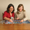 Two women smiling and working together on a jigsaw puzzle titled 'Rainy Day Stroll' by Cobble Hill, with scattered pieces on a wooden table. One woman holds a coffee mug, creating a cozy, collaborative atmosphere.