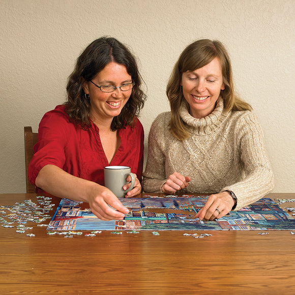 Two women smiling and working together on a jigsaw puzzle titled 'Rainy Day Stroll' by Cobble Hill, with scattered pieces on a wooden table. One woman holds a coffee mug, creating a cozy, collaborative atmosphere.