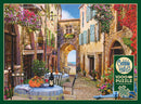 A charming jigsaw puzzle titled 'French Village' featuring a picturesque cobblestone street lined with quaint buildings adorned with colorful flowers. A table set for two with floral arrangements and a bottle of wine adds to the inviting atmosphere, leading to an archway that opens to a serene landscape. Created by Cobble Hill, this 1000-piece puzzle includes a poster.