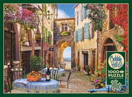 A charming jigsaw puzzle titled 'French Village' featuring a picturesque cobblestone street lined with quaint buildings adorned with colorful flowers. A table set for two with floral arrangements and a bottle of wine adds to the inviting atmosphere, leading to an archway that opens to a serene landscape. Created by Cobble Hill, this 1000-piece puzzle includes a poster.