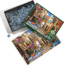 Cobble Hill jigsaw puzzle titled 'French Village' featuring a picturesque scene of a quaint European street adorned with colorful flowers, a cozy outdoor dining setup, and charming architecture. The puzzle contains 1000+ pieces and comes in an open box displaying the scattered pieces, with the artwork of the completed puzzle visible for reference.