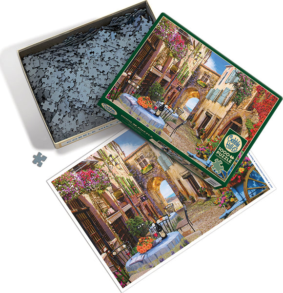 Cobble Hill jigsaw puzzle titled 'French Village' featuring a picturesque scene of a quaint European street adorned with colorful flowers, a cozy outdoor dining setup, and charming architecture. The puzzle contains 1000+ pieces and comes in an open box displaying the scattered pieces, with the artwork of the completed puzzle visible for reference.