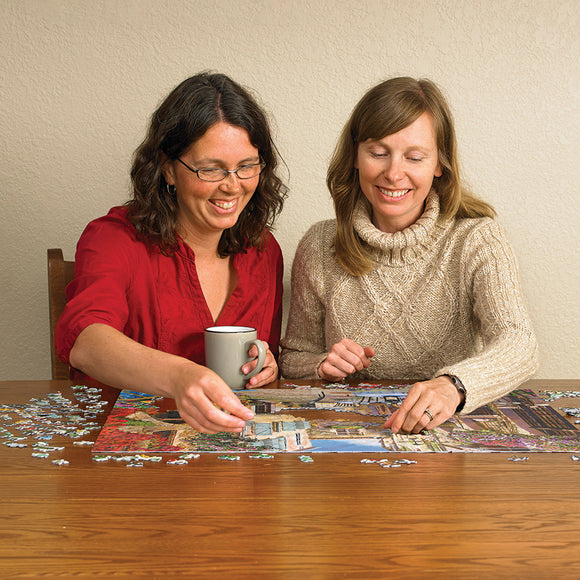 Two women joyfully working together on a colorful jigsaw puzzle titled 'French Village' by Cobble Hill. One woman is holding a cup, while they both concentrate on fitting pieces together on a wooden table, surrounded by scattered puzzle pieces.