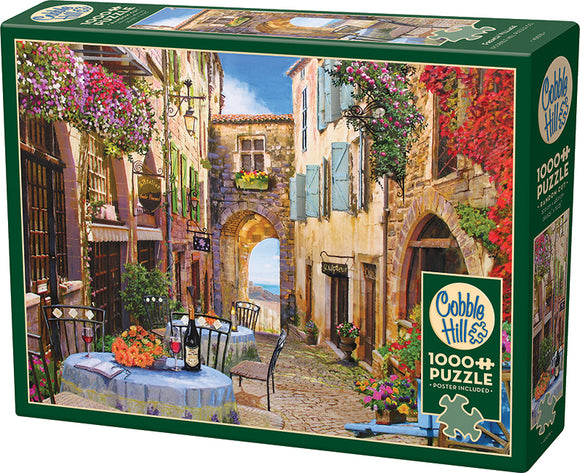 Cobble Hill French Village jigsaw puzzle featuring a quaint street scene. The artwork displays charming tables set with floral arrangements and wine bottles, framed by colorful buildings adorned with vibrant flowers. The puzzle contains 1000 pieces and includes a poster for reference.