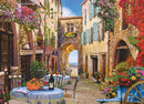 A picturesque jigsaw puzzle titled 'French Village' by Cobble Hill. The image depicts a charming cobblestone street in a French village adorned with colorful flowers, quaint buildings, and outdoor café tables. A bottle of wine and glasses sit on a table, inviting viewers to imagine a leisurely afternoon, while an archway frames a glimpse of the sea in the distance.