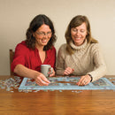 Two women smiling and working together on the Parisian Flowers jigsaw puzzle from Cobble Hill. One woman holds a cup while the other places a puzzle piece on the table filled with colorful pieces. The puzzle features vibrant flowers and scenic Parisian elements, showcasing a delightful puzzle experience.