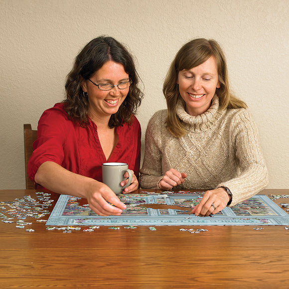 Two women smiling and working together on the Parisian Flowers jigsaw puzzle from Cobble Hill. One woman holds a cup while the other places a puzzle piece on the table filled with colorful pieces. The puzzle features vibrant flowers and scenic Parisian elements, showcasing a delightful puzzle experience.