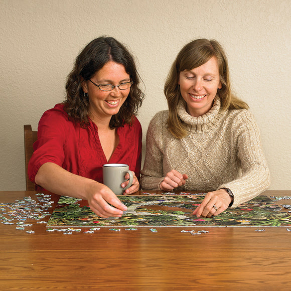 Two women enjoying a relaxing afternoon assembling the Cobble Hill Succulent Garden jigsaw puzzle. One woman holds a mug while reaching for a puzzle piece, and the table is strewn with colorful puzzle pieces and the nearly completed vibrant succulent garden image.